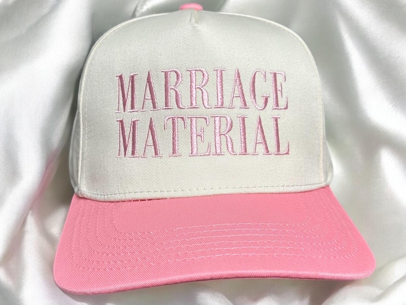 May include: A white baseball cap with a pink brim. The cap has the words "Marriage Material" embroidered in pink on the front.