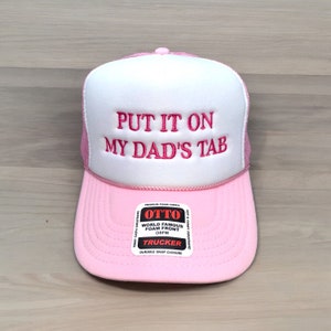 May include: Pink trucker hat with a white front panel and pink mesh back. The text "PUT IT ON MY DAD'S TAB" is embroidered in pink. The hat features a snap closure and a "OTTO" brand label.