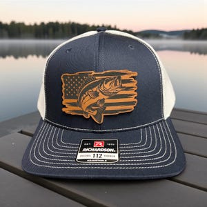May include: Navy blue and white trucker hat with a leather patch featuring a fish leaping over a distressed American flag. The hat has a curved brim, white mesh back, and adjustable strap. The patch reads "RICHARDSON ORIGINAL 112 TRUCKER".