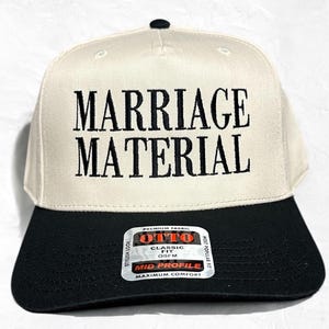 May include: A white baseball cap with black embroidery that reads "Marriage Material". The cap has a black brim and a black label on the back that reads "Otto Classic Fit OSFM Mid Profile Maximum Comfort".