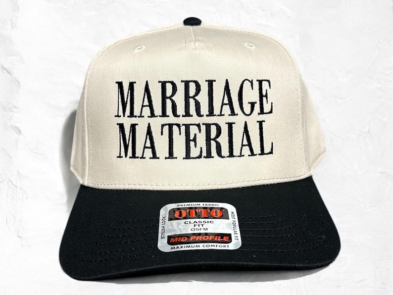 May include: A white baseball cap with black embroidery that reads "Marriage Material". The cap has a black brim and a black label on the back that reads "Otto Classic Fit OSFM Mid Profile Maximum Comfort".