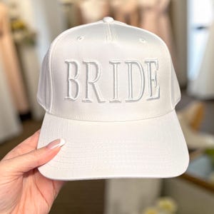 May include: A white baseball cap with the word "BRIDE" embroidered in capital letters across the front. The hat has a curved brim and a rounded top. The hat is made of a smooth, possibly satin-like material.