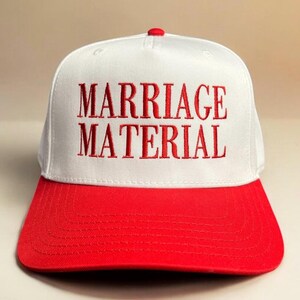 May include: White baseball cap with a red brim. The front of the cap has the words "MARRIAGE MATERIAL" embroidered in red.