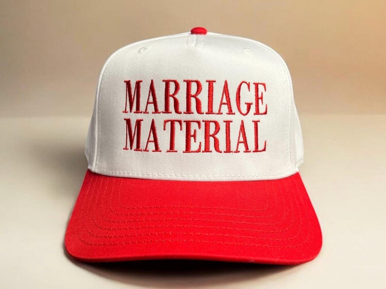 May include: White baseball cap with a red brim. The front of the cap has the words "MARRIAGE MATERIAL" embroidered in red.
