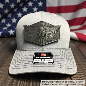 May include: Heather grey and black trucker hat with a vintage dark grey patch. The patch features an eagle, the text "250 YEARS", "1776", "MADE IN AMERICA", and "2026". The hat has a curved brim and an adjustable strap.