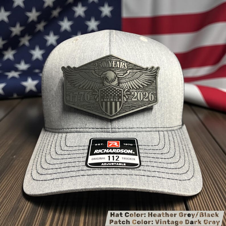 May include: Heather grey and black trucker hat with a vintage dark grey patch. The patch features an eagle, the text "250 YEARS", "1776", "MADE IN AMERICA", and "2026". The hat has a curved brim and an adjustable strap.