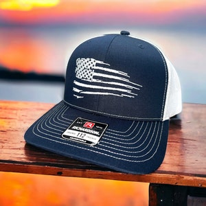 May include: A navy blue and white trucker hat with a white distressed American flag design. The hat has a white mesh back and a black adjustable strap with a red label that says "Richardson 112 Original Trucker Adjustable".
