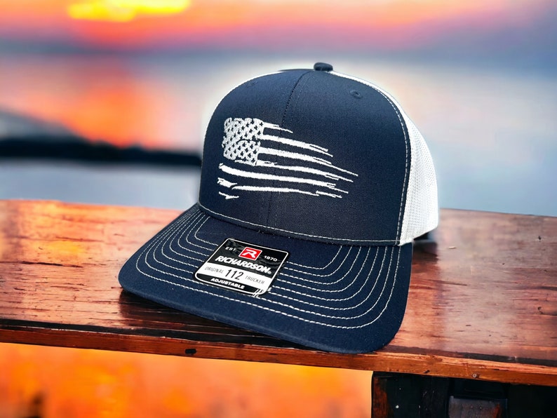 May include: A navy blue and white trucker hat with a white distressed American flag design. The hat has a white mesh back and a black adjustable strap with a red label that says "Richardson 112 Original Trucker Adjustable".