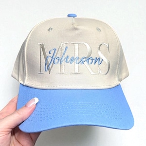 Peut inclure: Casquette de baseball beige et bleu clair avec le mot "MRS" et le nom "Johnson" brodés en fil bleu clair. La casquette a une visière incurvée et un bouton sur le dessus.