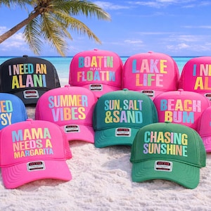 May include: A collection of colorful trucker hats with various phrases like "I Need An Alani", "Summer Vibes", and "Chasing Sunshine". The hats are displayed on a sandy beach with a palm tree and ocean in the background.