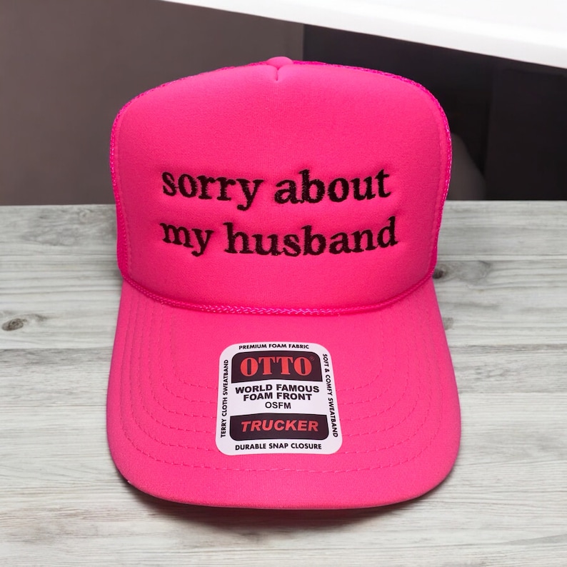 May include: Pink trucker hat with a black embroidered message that says "sorry about my husband". The hat has a black and white label that says "OTTO World Famous Foam Front OSFM Trucker Durable Snap Closure".