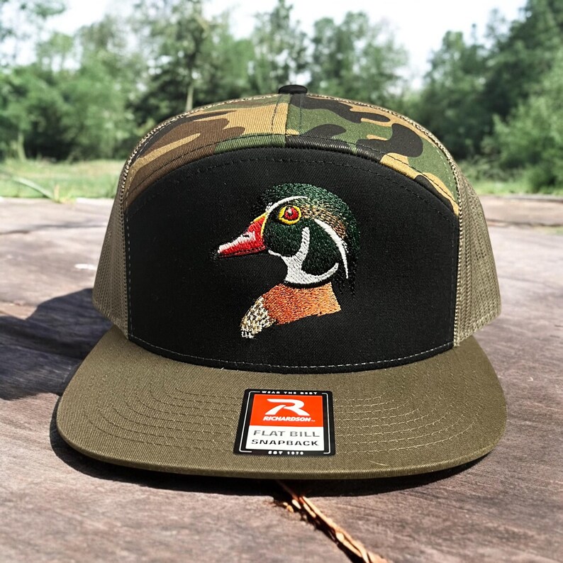 May include: A green, black and brown camouflage flat bill snapback hat with an embroidered wood duck design. The hat has a Richardson brand label on the front.