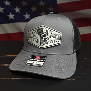 K&ouml;nnte beinhalten: Graue Trucker-Kappe mit schwarzem Mesh-R&uuml;cken. Auf der Vorderseite befindet sich ein wei&szlig;er Aufn&auml;her mit einem Totenkopf, der amerikanischen Flagge und dem Text "WE THE PEOPLE 1776". Die Kappe hat einen gebogenen Schirm und einen verstellbaren Riemen.