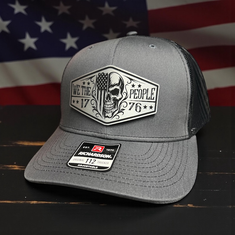 K&ouml;nnte beinhalten: Graue Trucker-Kappe mit schwarzem Mesh-R&uuml;cken. Auf der Vorderseite befindet sich ein wei&szlig;er Aufn&auml;her mit einem Totenkopf, der amerikanischen Flagge und dem Text "WE THE PEOPLE 1776". Die Kappe hat einen gebogenen Schirm und einen verstellbaren Riemen.