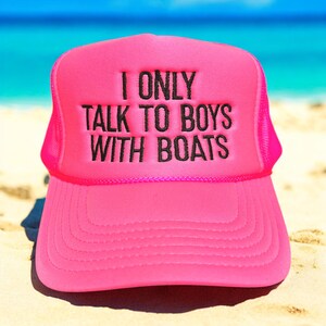 May include: Bright pink trucker hat with the text "I ONLY TALK TO BOYS WITH BOATS" in black. The hat has a mesh back and a curved brim. The hat is sitting on a sandy surface with a blue ocean in the background.