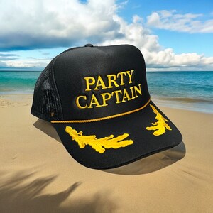 May include: Black trucker hat with a yellow mesh back and a gold embroidered rope trim. The hat has the words "PARTY CAPTAIN" embroidered in yellow on the front.