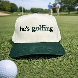 May include: A white and green baseball cap with the text "he's golfing" embroidered in green. The cap is on a green golf course, with a golf ball in the foreground and golfers in the background.