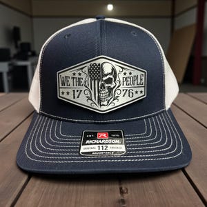 K&ouml;nnte beinhalten: Marineblaue und wei&szlig;e Trucker-Cap mit einem Aufn&auml;her mit einem Totenkopf, der amerikanischen Flagge und dem Text "WE THE PEOPLE 1776". Die Cap hat einen gebogenen Schirm und einen verstellbaren Riemen. Marke Richardson.