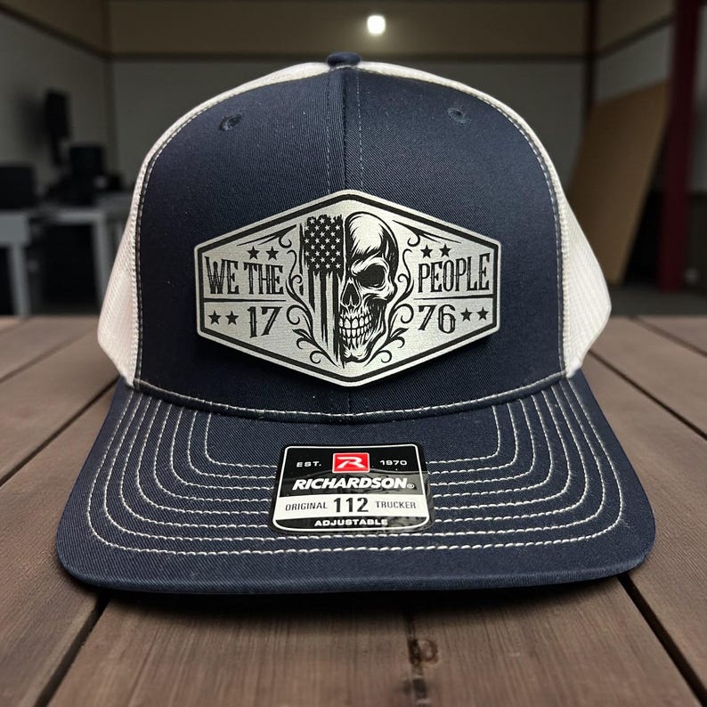 K&ouml;nnte beinhalten: Marineblaue und wei&szlig;e Trucker-Cap mit einem Aufn&auml;her mit einem Totenkopf, der amerikanischen Flagge und dem Text "WE THE PEOPLE 1776". Die Cap hat einen gebogenen Schirm und einen verstellbaren Riemen. Marke Richardson.