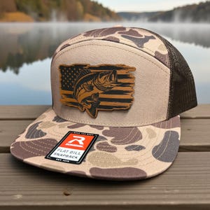 May include: A flat bill snapback hat with a camouflage pattern and a brown mesh back. The hat features a wooden patch with a fish and American flag design. The tag reads "FLAT BILL SNAPBACK".