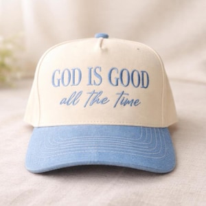 May include: A cream-colored baseball cap featuring the embroidered text GOD IS GOOD all The Time in light blue thread. The cap has a light blue brim with visible stitching and a structured crown with a small button at the top center.