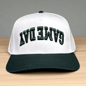 May include: A white baseball cap with a dark green brim and dark green embroidered text on the front that reads GAME DAY in an upside-down orientation. The cap features a structured crown with a top button and stitched detailing on the brim.