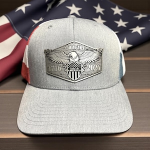 May include: Gray trucker hat with a silver emblem featuring an eagle, shield, and the text "250 YEARS AMERICA 1776-2026." The hat has a curved brim and a mesh back. An American flag is in the background.
