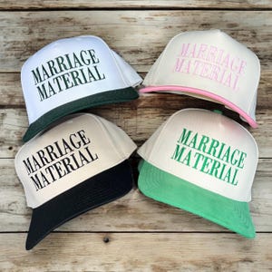 May include: Four white baseball caps with a black, green, or pink brim. Each cap has the text "Marriage Material" embroidered in green or pink letters.