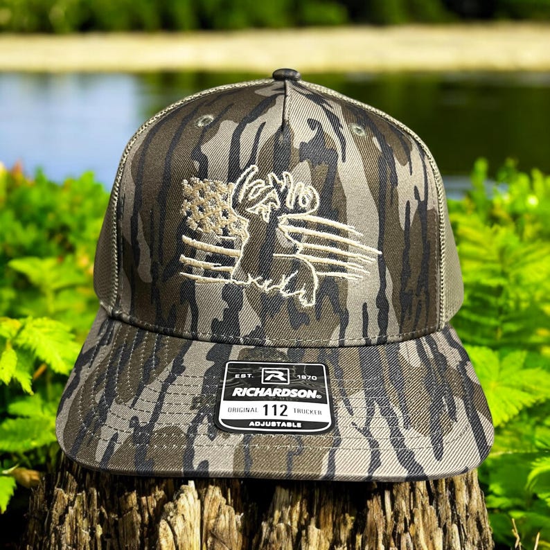 May include: A camouflage baseball cap with a brown and white embroidered American flag and a deer silhouette. The cap has a black mesh back and a black adjustable strap with the text "Richardson. Original Trucker 112 Adjustable" on a white label.