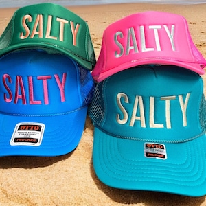 May include: Four trucker hats with mesh backs and foam fronts, each featuring the word SALTY in block letters. The hats are green, bright blue, neon pink, and teal. Two hats have Otto brand labels on the brims. The hats are stacked together.