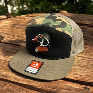 May include: A green, black and camouflage flat bill snapback hat with a wood duck embroidered on the front. The hat has a mesh back and a "Remarkable" label on the side.