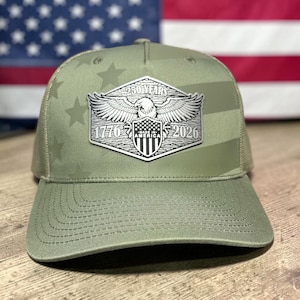 May include: Olive green trucker hat with a mesh back and a curved brim. The front features a silver emblem with an eagle, shield, and the text "250 YEARS 1776-2026 AMERICA". The hat has a star pattern on the front panel.