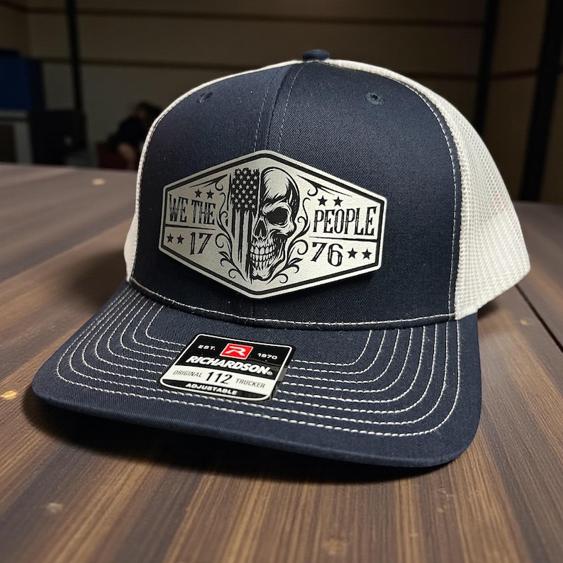 K&ouml;nnte beinhalten: Marineblaue Trucker-M&uuml;tze mit wei&szlig;em Mesh-R&uuml;cken und gebogenem Schirm. Auf der Vorderseite befindet sich ein Aufn&auml;her mit einem Totenkopf, der amerikanischen Flagge und dem Text "WE THE PEOPLE 17 76". Die M&uuml;tze ist verstellbar.