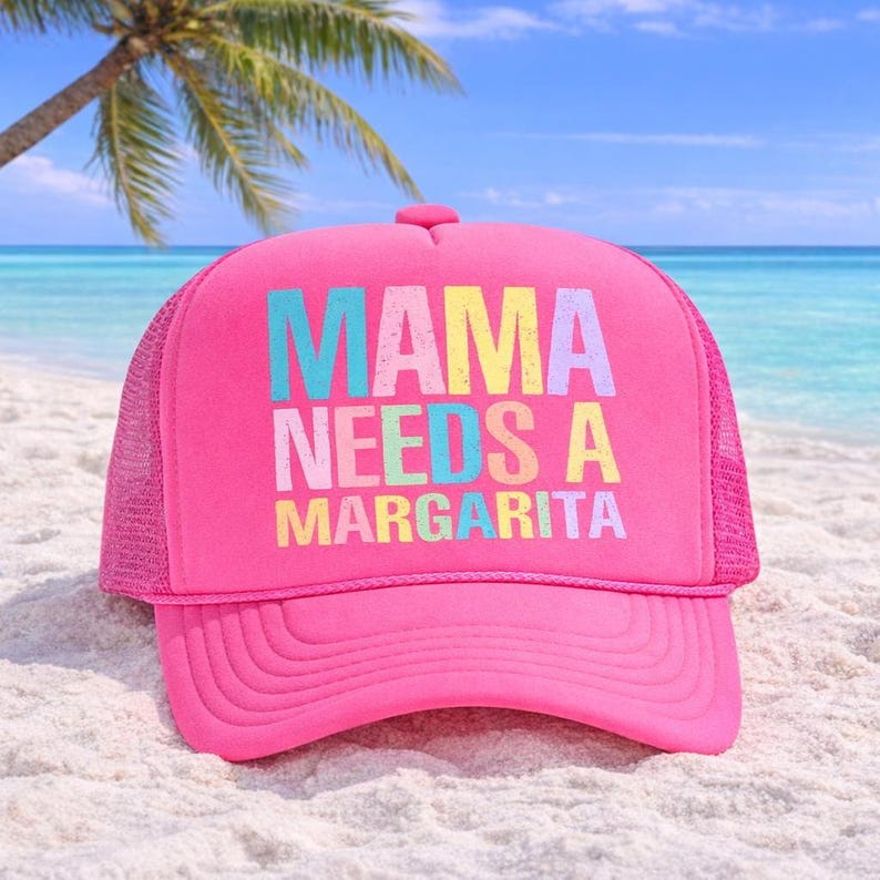 May include: A bright pink trucker hat with the text "MAMA NEEDS A MARGARITA" in colorful block letters. The hat is on a sandy beach with a palm tree and ocean in the background.