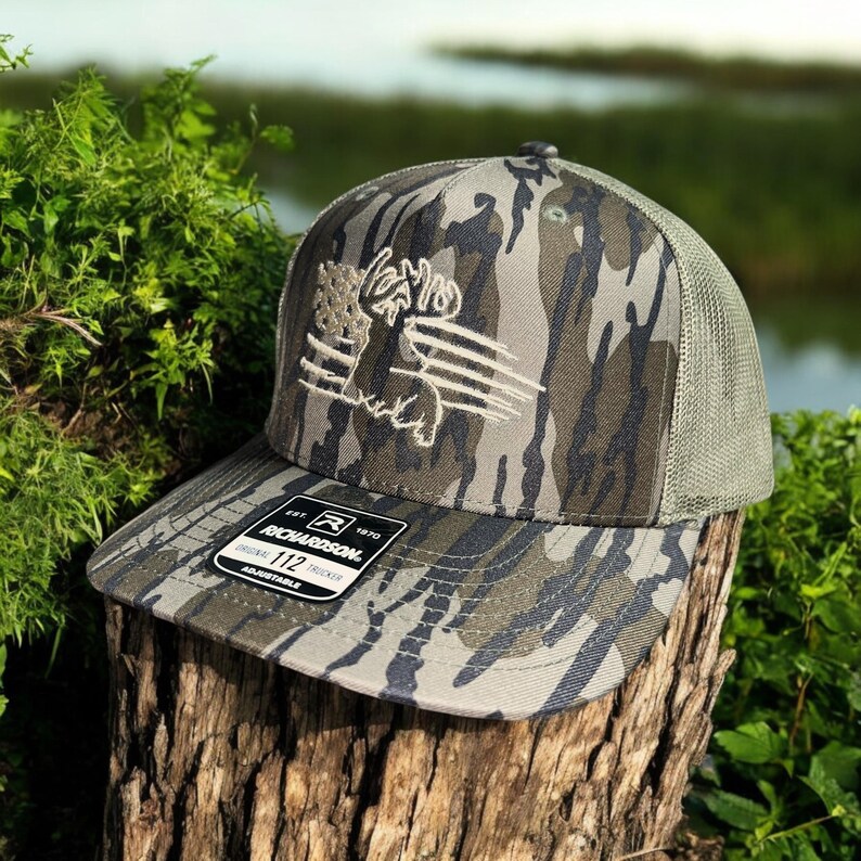May include: A camouflage trucker hat with a white embroidered moose logo and the text "Richardson 112 Original Trucker Adjustable" on the front.