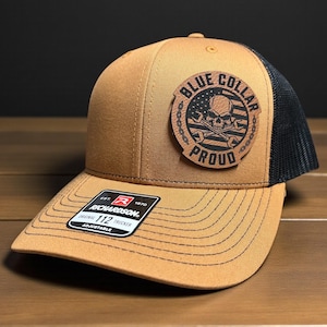 May include: A tan and black trucker cap featuring a patch with the words "Blue Collar Proud", a skull, and crossed wrenches. The cap has a curved peak and an adjustable strap. The Richardson brand label is visible.