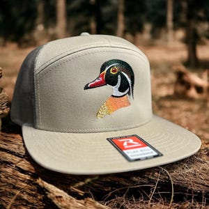 May include: A beige and black baseball cap with a wood duck embroidered on the front. The cap has a mesh back and a flat bill.