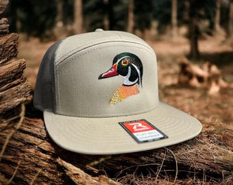 Op de afbeelding: Een beige en zwarte baseballcap met een gebrodeerde houtentekening op de voorkant. De cap heeft een mesh achterkant en een platte klep.