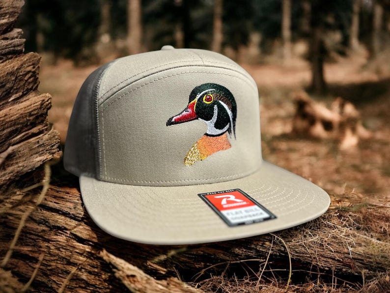 May include: A beige and black baseball cap with a wood duck embroidered on the front. The cap has a mesh back and a flat bill.