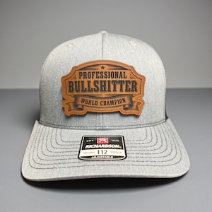 May include: A gray trucker hat with a brown leather patch that reads "PROFESSIONAL BULLSHITTER WORLD CHAMPION." The hat has a curved brim and an adjustable strap. The Richardson brand tag is visible.