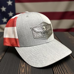 May include: A gray baseball cap with a mesh back and a red, white, and red pattern on the side. The front features a silver emblem with an eagle, the text "250 YEARS", "1776", and "MADE IN AMERICA".