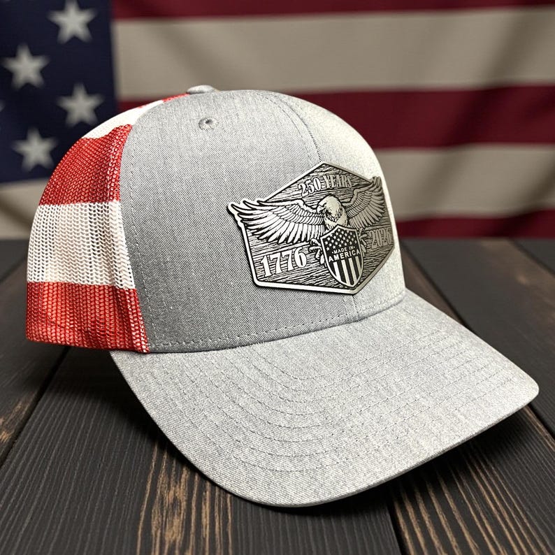 May include: A gray baseball cap with a mesh back and a red, white, and red pattern on the side. The front features a silver emblem with an eagle, the text "250 YEARS", "1776", and "MADE IN AMERICA".