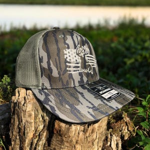 May include: A camouflage trucker hat with a white embroidered American flag and a deer head design. The hat has a mesh back and a black adjustable strap with the text "RECHARGED" and "112" on it.
