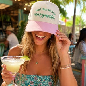 May include: A baseball cap with a white crown and a pink peak, featuring the text "bad day to be a margarita" in green. A margarita cocktail with a lime garnish is held, with tortilla chips and salsa on the table.