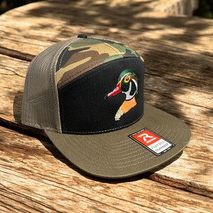 May include: A black and green camouflage trucker hat with a black crown and a green mesh back. The hat features an embroidered wood duck design on the front. The hat has a flat bill and a red "R" logo on the back.