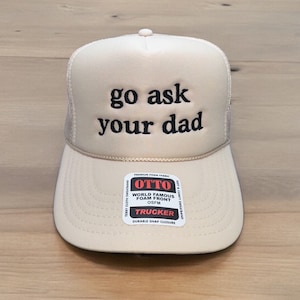 May include: A beige trucker hat with the phrase "go ask your dad" embroidered in black. The hat features a mesh back, a curved brim, and a snap closure. A small label reads "OTTO TRUCKER".