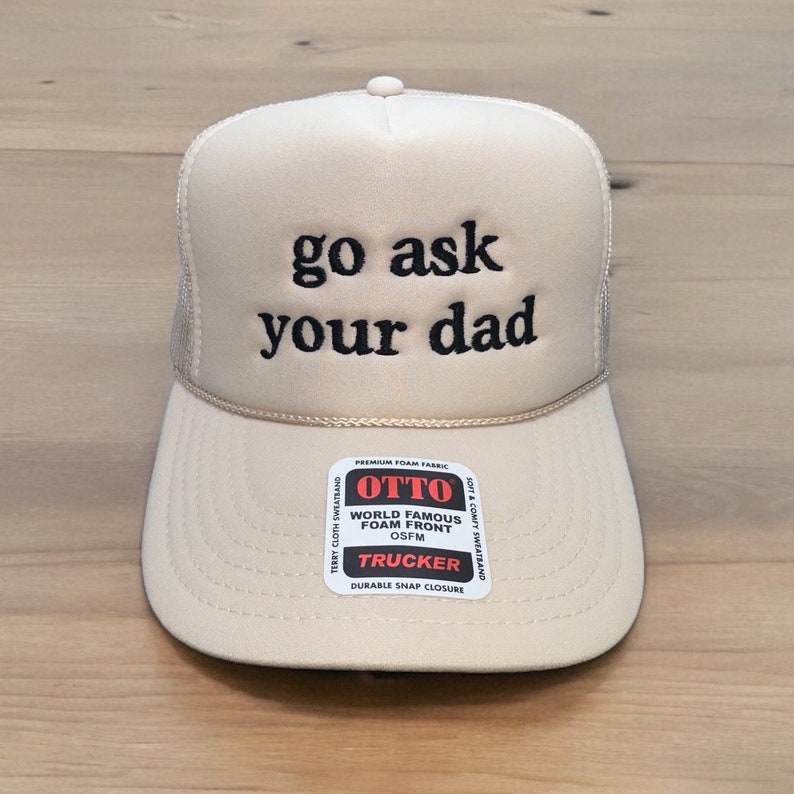 May include: A beige trucker hat with the phrase "go ask your dad" embroidered in black. The hat features a mesh back, a curved brim, and a snap closure. A small label reads "OTTO TRUCKER".