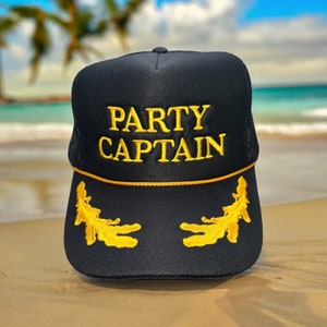 May include: Black mesh trucker hat with gold embroidery that reads "PARTY CAPTAIN". The hat has two gold embroidered leaf designs on the sides.