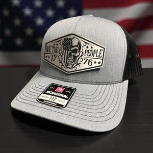 K&ouml;nnte beinhalten: Graue Trucker-Cap mit schwarzem Mesh-R&uuml;cken. Auf der Vorderseite befindet sich ein silberner Aufn&auml;her mit Totenkopf, amerikanischer Flagge und dem Text "WE THE PEOPLE 1776". Mit gebogenem Schirm und verstellbarem Riemen.