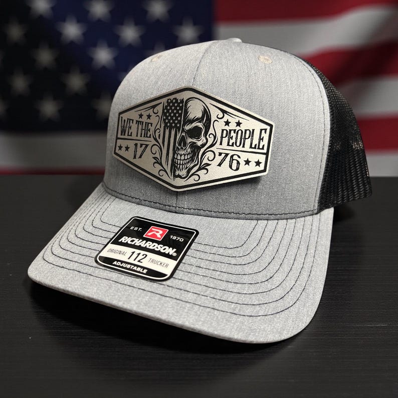 K&ouml;nnte beinhalten: Graue Trucker-Cap mit schwarzem Mesh-R&uuml;cken. Auf der Vorderseite befindet sich ein silberner Aufn&auml;her mit Totenkopf, amerikanischer Flagge und dem Text "WE THE PEOPLE 1776". Mit gebogenem Schirm und verstellbarem Riemen.